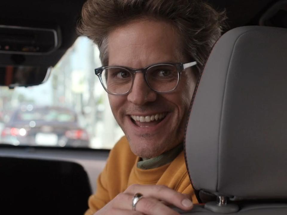 Ryan Piers Williams in barbie as gloria's husband, smiling from the front seat of a car