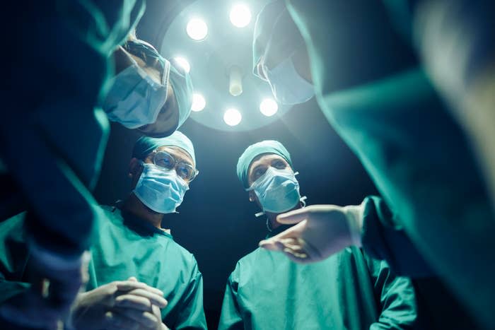 Surgeons wearing blue scrubs, masks, and gloves gathered under an operating room light, seemingly in the middle of a surgery or medical procedure