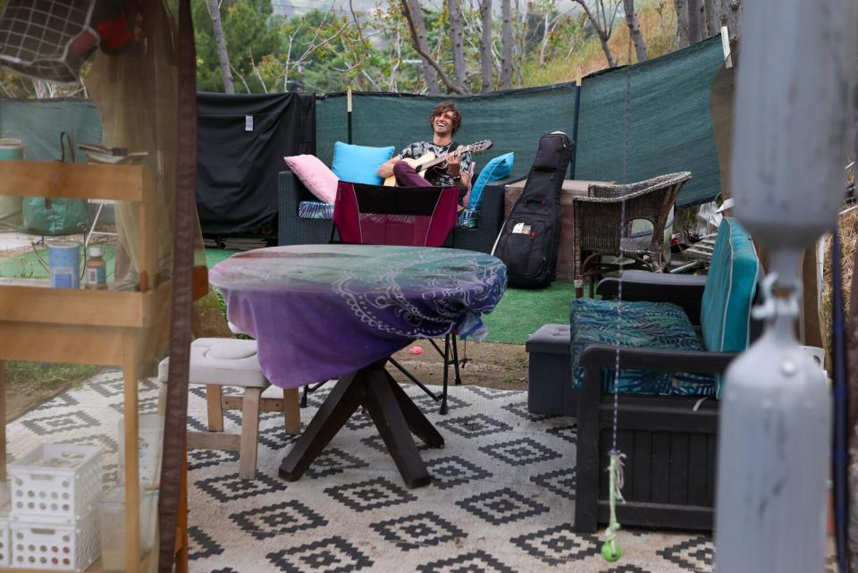 A seated man holds a guitar in a furnished yard.