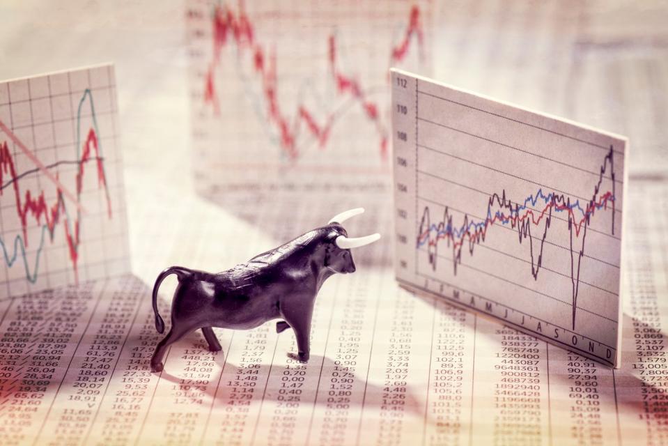 A bull figurine placed in front of three volatile stock charts that have risen up from a financial newspaper. 