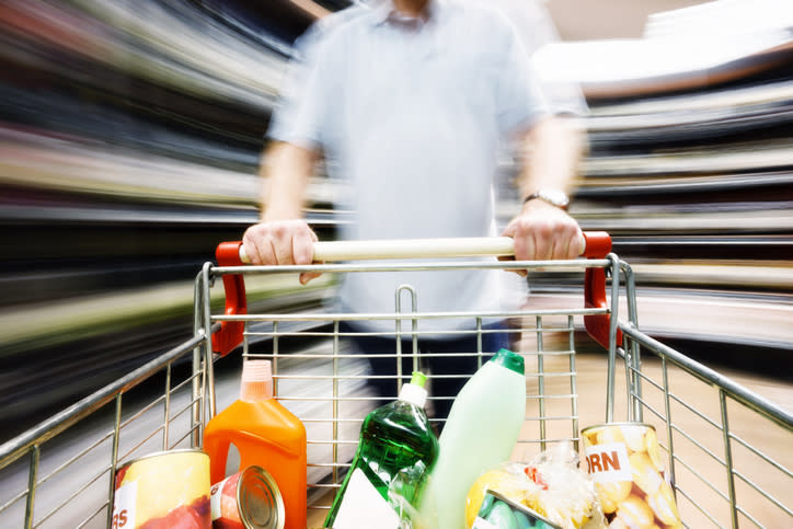 A blurred image of a person pushing a shopping cart filled with various groceries in a supermarket aisle. The person's identity is not visible