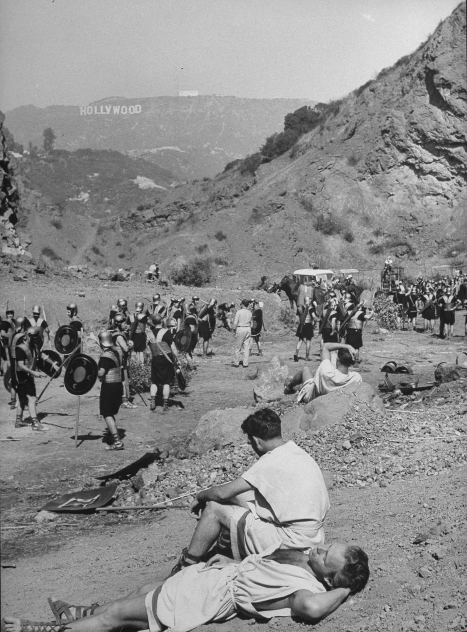 Extras portraying Roman soldiers resting in treeless valley, with Hollywood sign visible on hill in background during filming of Julius Caesar.