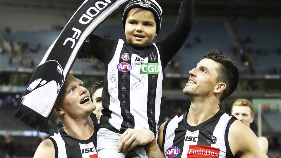 Collingwood mascot, Kyron McGuire, smiles and is carried off the ground by Adam Treloar and Scott Pendlebury.