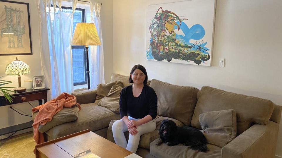 A woman sits on a sofa with her dog inside an apartment