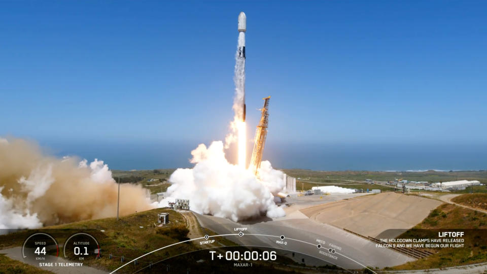 a black and white spacex falcon 9 rocket launches into a blue sky with the ocean in the background