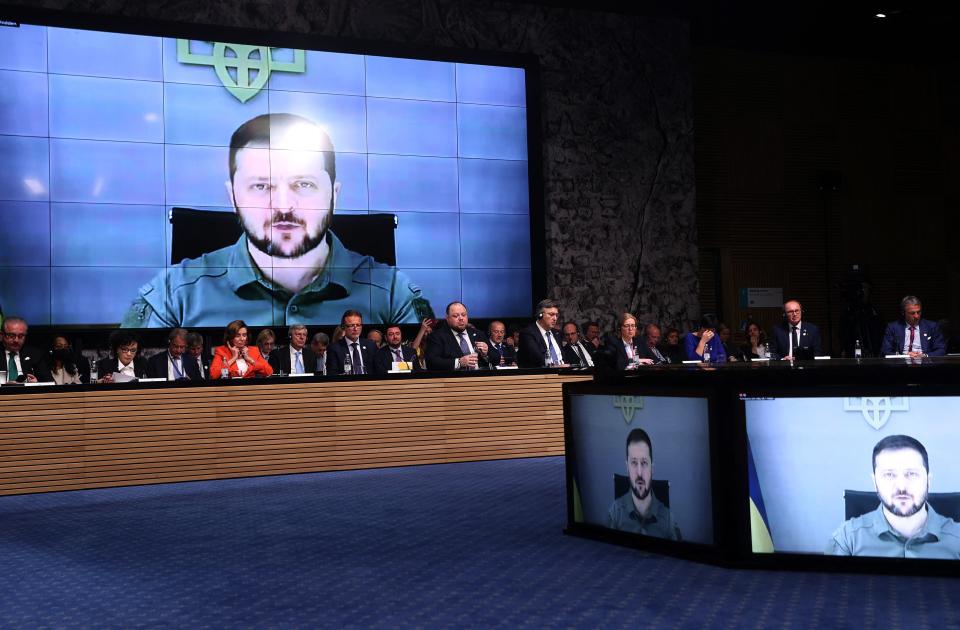 President Volodymyr Zelensky appears on a large video screen above a panel of conference participants, with two other screens carrying the image in the foreground.