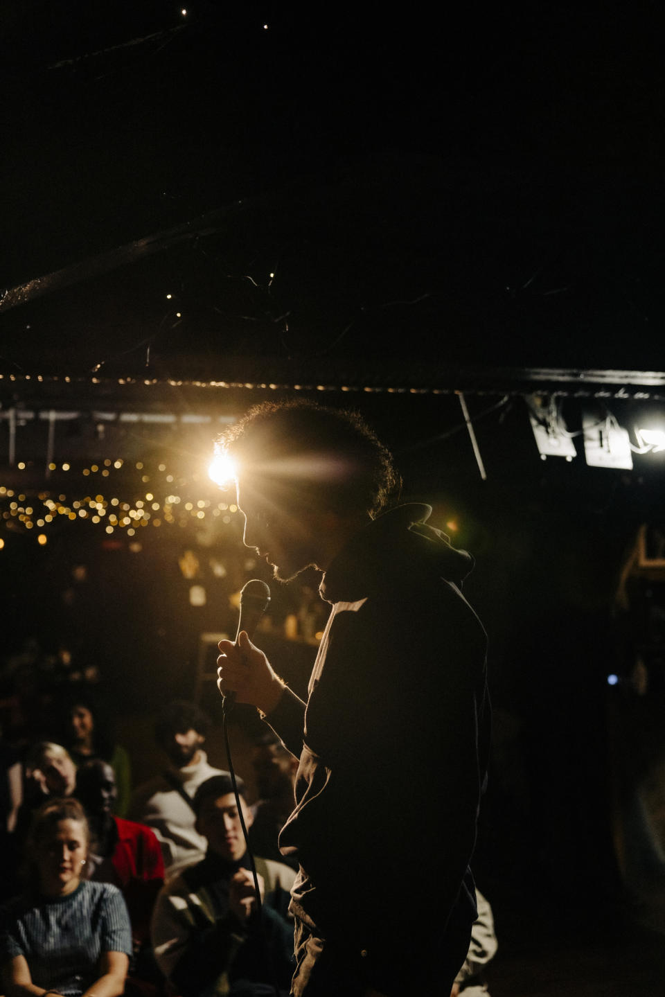 Man in a hoodie performing stand-up comedy in front of a small audience; string lights hang in the background