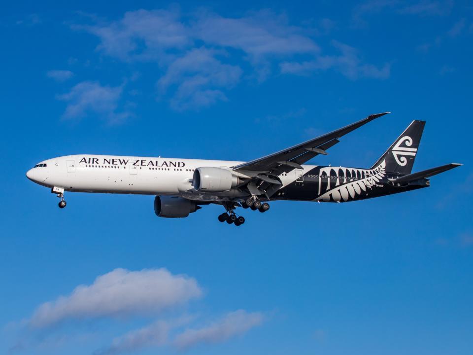 An Air New Zealand Boeing 777 flying in a lightly cloudy sky with its landing gear down.