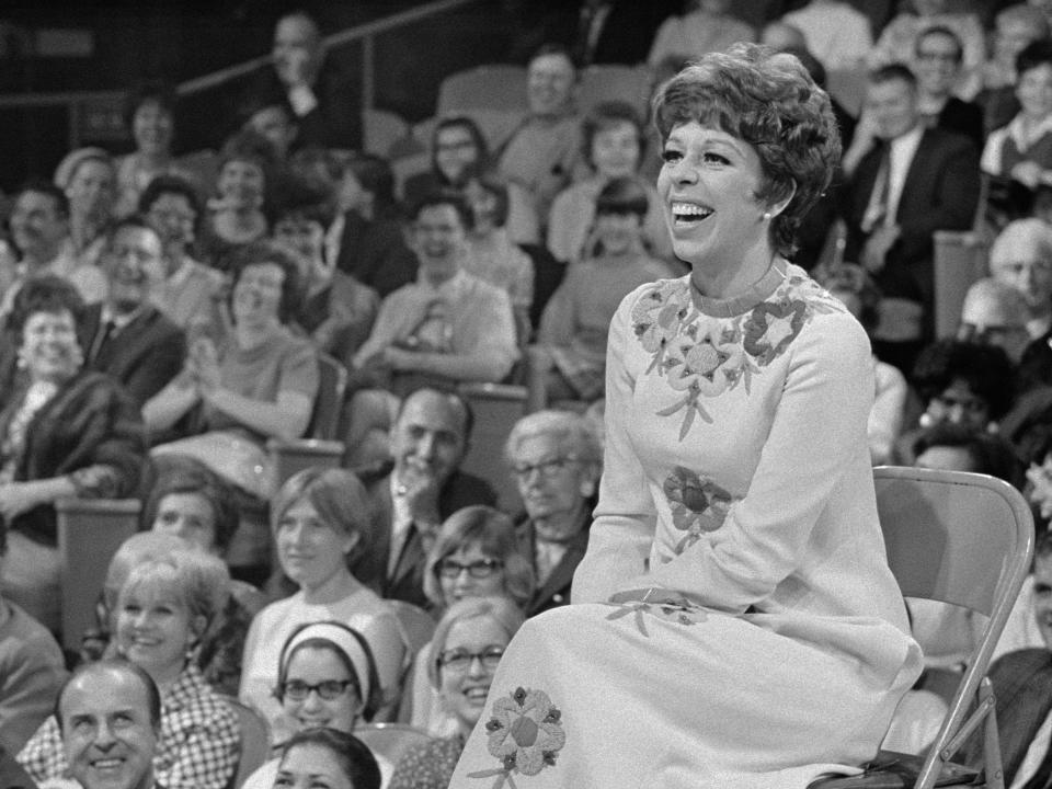 Black and white photo of Carol Burnett sitting on stage surrounded by a laughing audience.