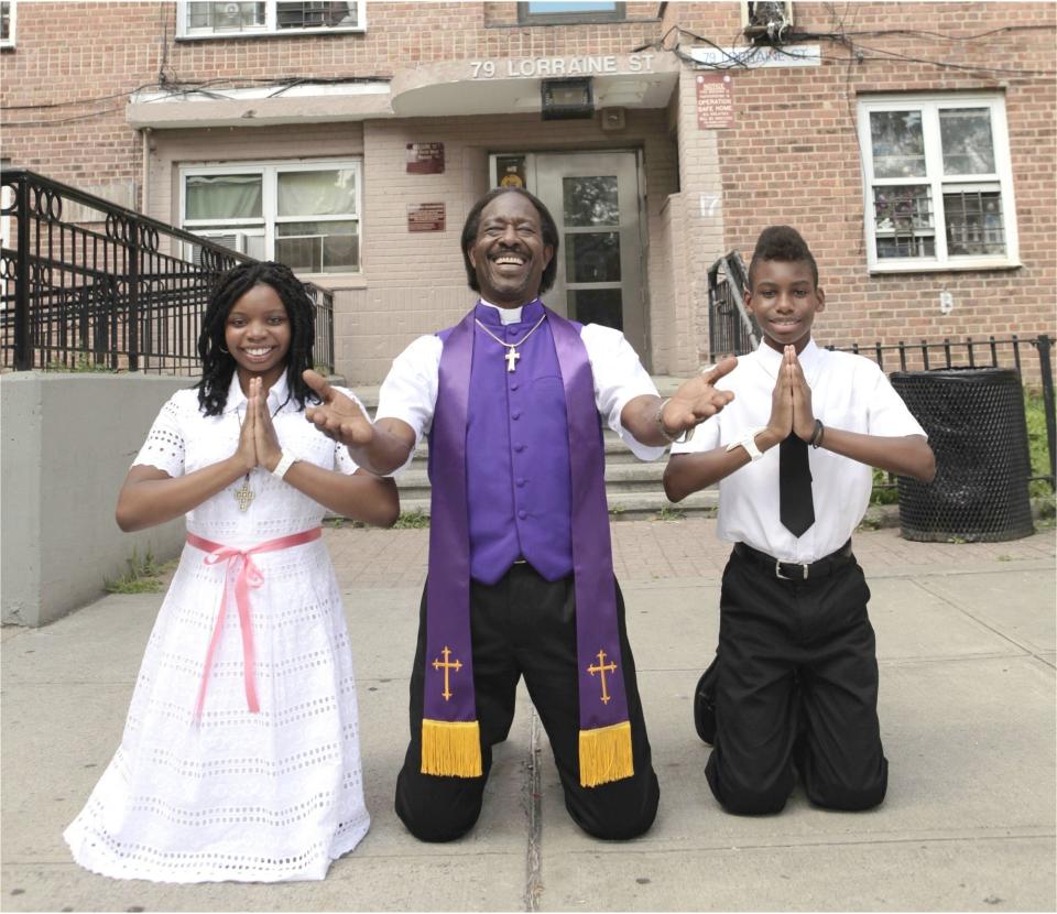 Two children kneel in praying positions on either side of a smiling pastor