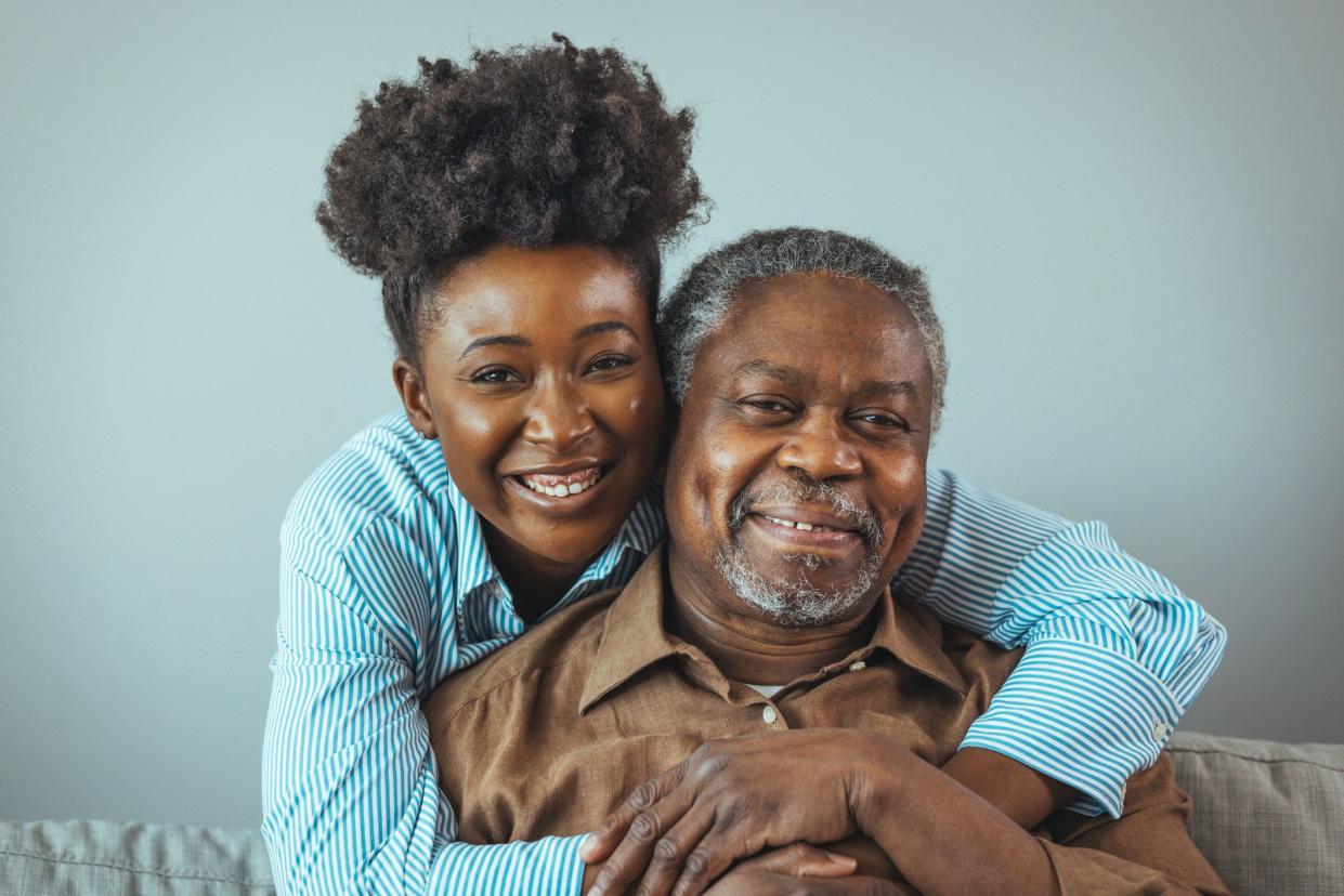 Senior man and his middle aged daughter smiling at each other embracing, close up. Portrait of a daughter holding her elderly father, sitting on a bed by a window in her father's room.