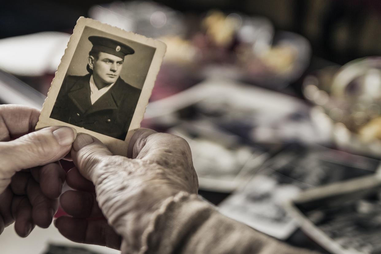 Focus on old woman's hand holding a vintage black and white portrait photograph of a young man in a military uniform with a blurred background of a pile of vintage photographs
