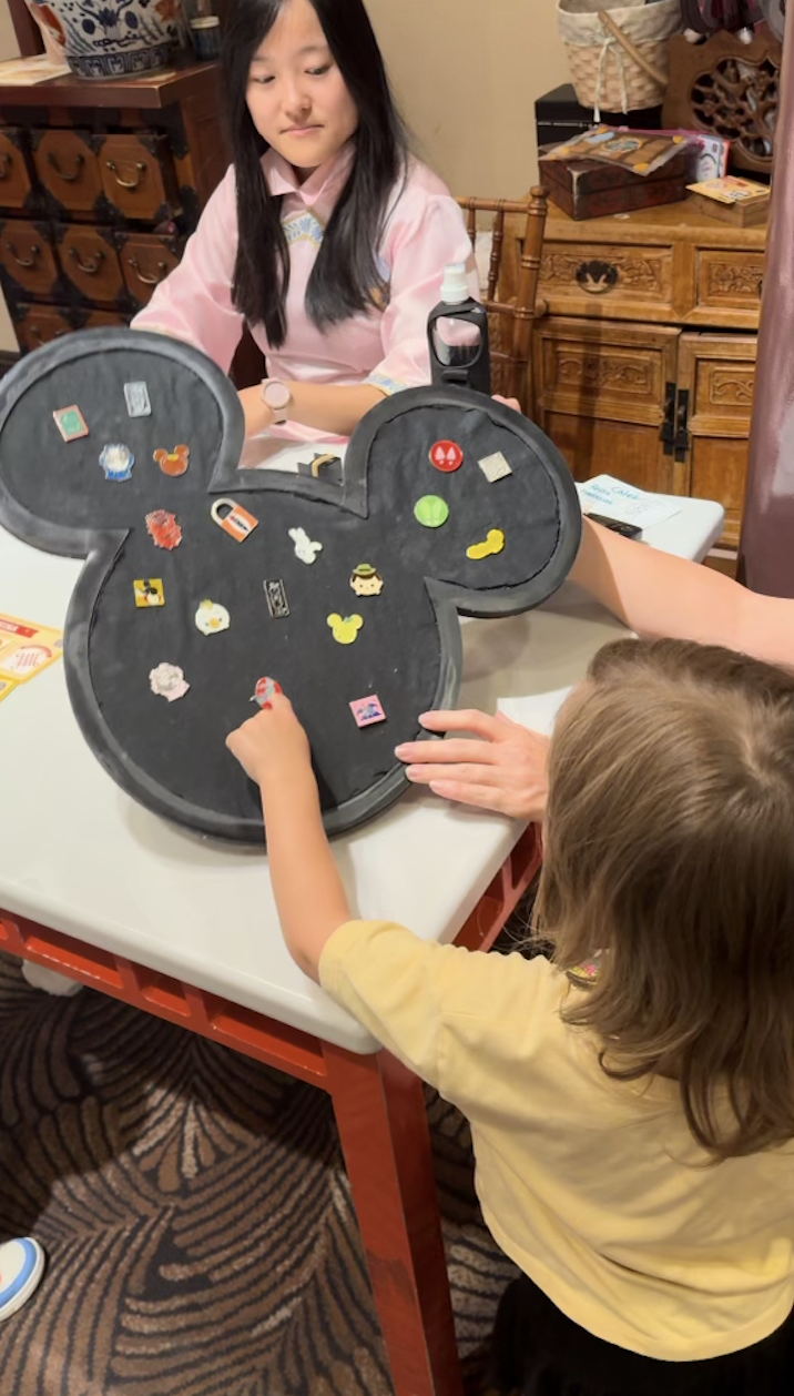 A young girl interacts with a Mickey-shaped board featuring various Disney-themed pins. An adult woman looks on from across the table