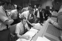In this April 15, 1970 photo made available by NASA, a group of flight controllers gather around the console of Glenn S. Lunney, foreground seated, Shift 4 flight director, in the Mission Operations Control Room (MOCR) of Mission Control Center (MCC) in Houston. Their attention is drawn to a weather map of the proposed landing site in the Pacific Ocean. At this point, the Apollo 13 lunar landing mission had been canceled, and the problem-plagued Apollo 13 crew members were in trans-Earth trajectory attempting to bring their crippled spacecraft back home. (NASA via AP)