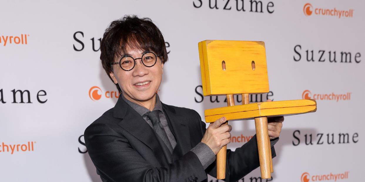 makoto shinkai, a middle-aged japanese man, smiling on the red carpet for his newest film suzume. shinkai wears round glasses and a black suit with a grey shirt, and he's holding up a three legged children's chair that references an object in the film