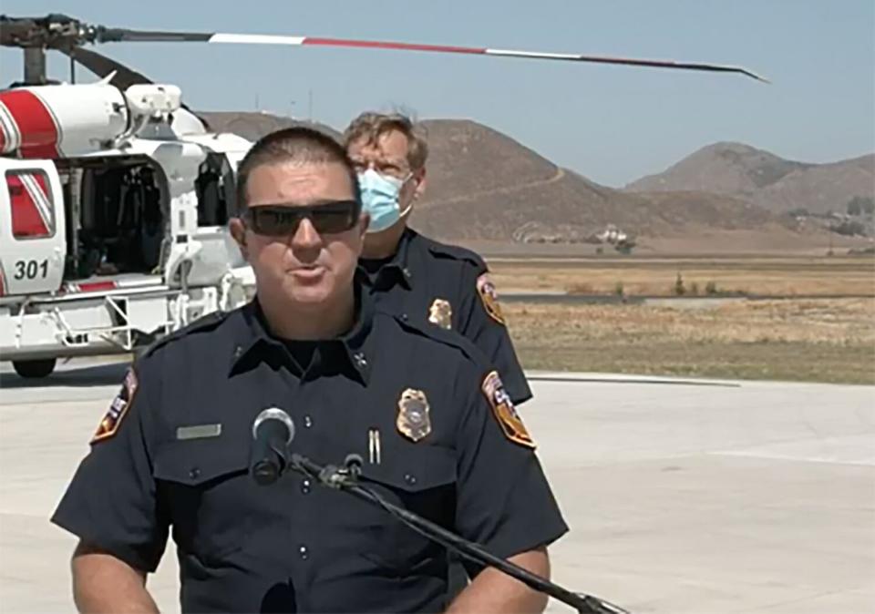 A man with short hair, in sunglasses and a dark uniform, stands before a mic. Behind him are another man and a helicopter.