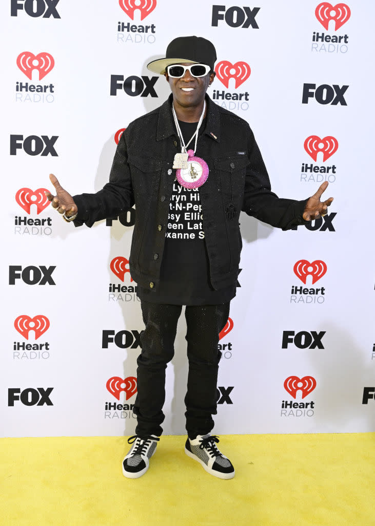 Person in a black denim jacket and pants with sneakers posing with hands out on the iHeartRadio event backdrop