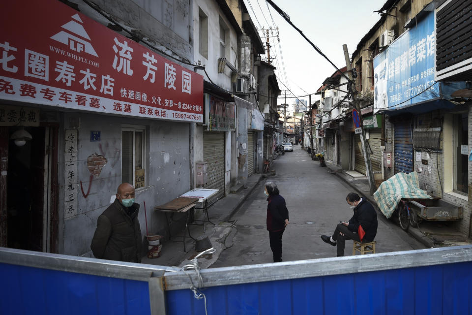 In this Sunday, Feb. 23, 2020, photo, a volunteer sits on a chair keep guard as residents stand near the barricades blocked a residential area in Wuhan in central China's Hubei province Sunday. Warning that China's virus epidemic is "still grim and complex," President Xi Jinping called Sunday for more efforts to stop the outbreak, revive industry and prevent the disease from disrupting spring planting of crops. (Chinatopix via AP)