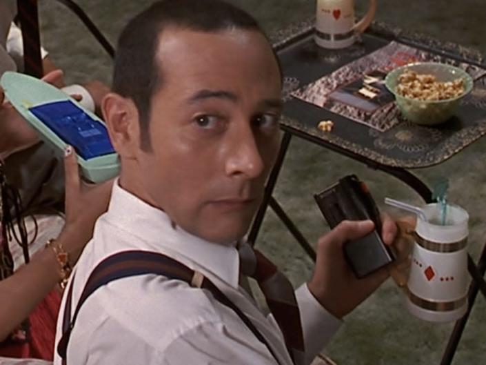 Paul Reubens as an FBI agent wearing a white shirt and suspenders and holding a coffee mug sitting on a couch and looking behind him