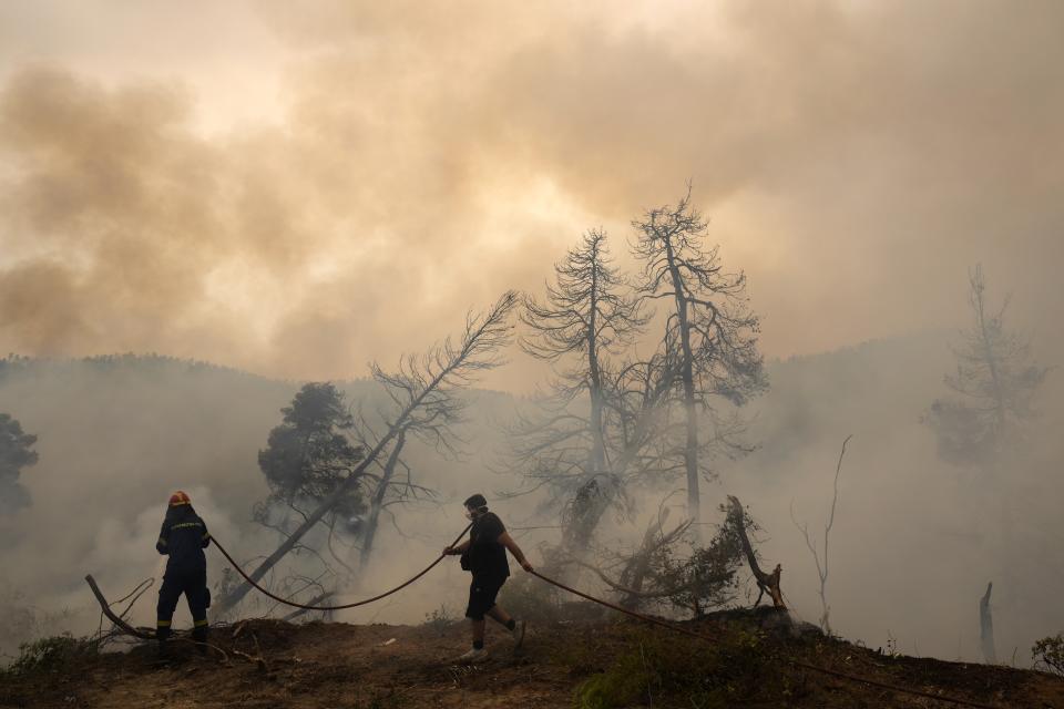 A firefighter tries to extinguish the flames as a local resident holds a water hose during a wildfire at Ellinika village on Evia island, about 176 kilometers (110 miles) north of Athens, Greece, Monday, Aug. 9, 2021. Firefighters and residents battled a massive forest fire on Greece's second largest island for a seventh day Monday, fighting to save what they can from flames that have decimated vast tracts of pristine forest, destroyed homes and businesses and sent thousands fleeing. (AP Photo/Petros Karadjias)