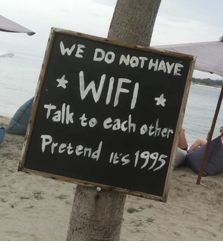 Chalkboard sign on a beach reads "WE DO NOT HAVE WIFI talk to each other Pretend it's 1995."