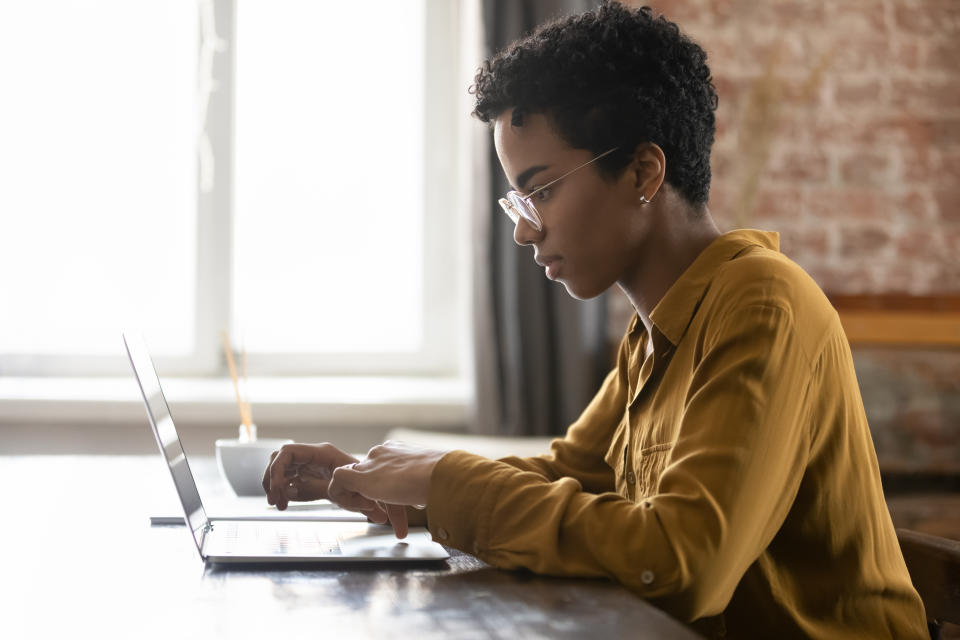 Side view focused young African American businesswoman in eyewear working on computer, sitting at table in modern loft office room, typing message, preparing electronic document or presentation.