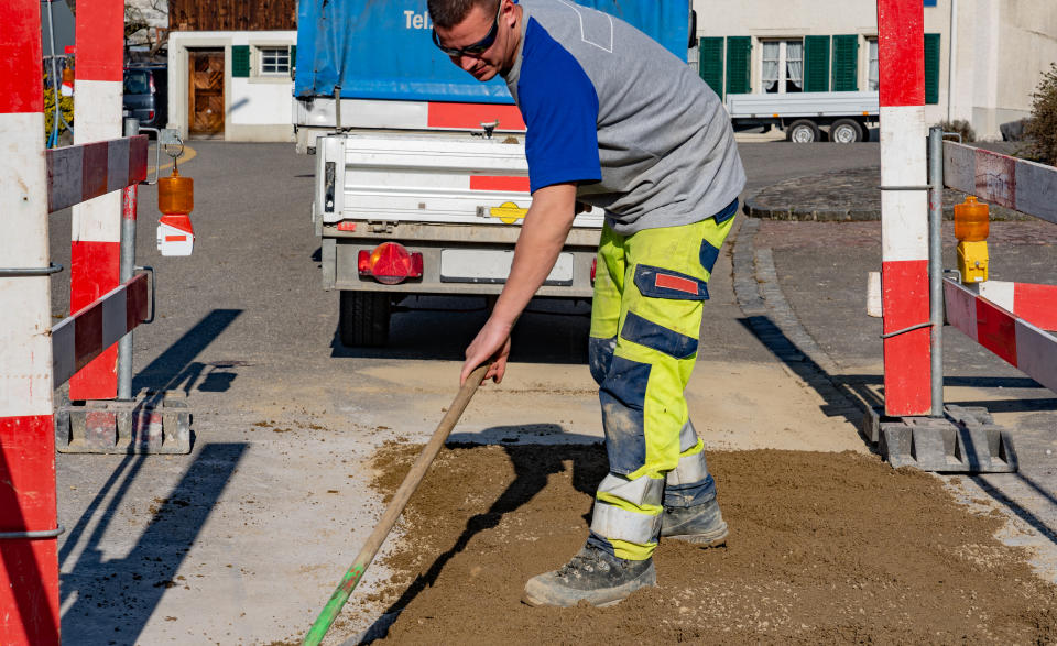 Construction worker with sunglasses and broomstick scrubbing sand in construction site in front of a construction vehicle and trailer. concept, close-up of worker.