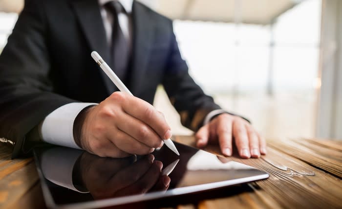 Man in suit viewed from neck-down signs an electronic tablet with a stylus.