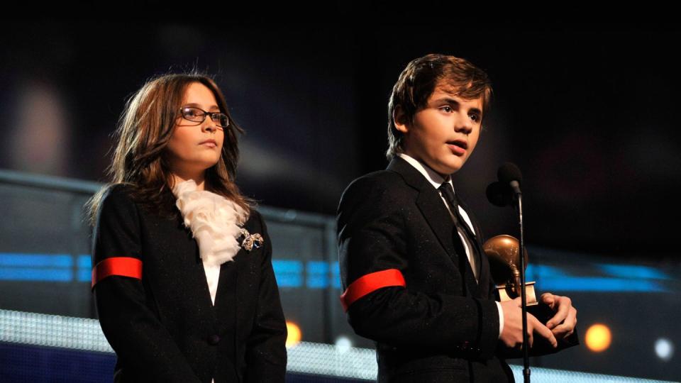 paris jackson stands with her hands clasp on a stage, prince jackson speaks into a microphone to the right while holding a grammy award, both children wear black suits with a red arm band on one sleeve and white dress shirts