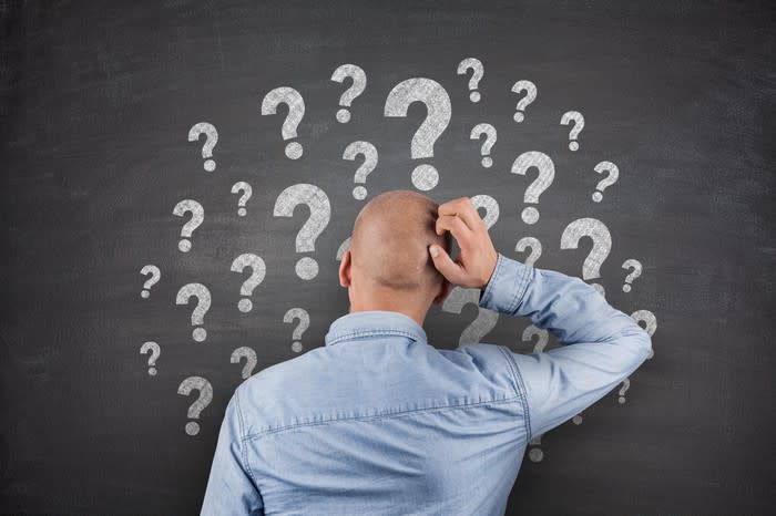 Man looking at chalkboard with many chalk question marks while scratching his bald head.