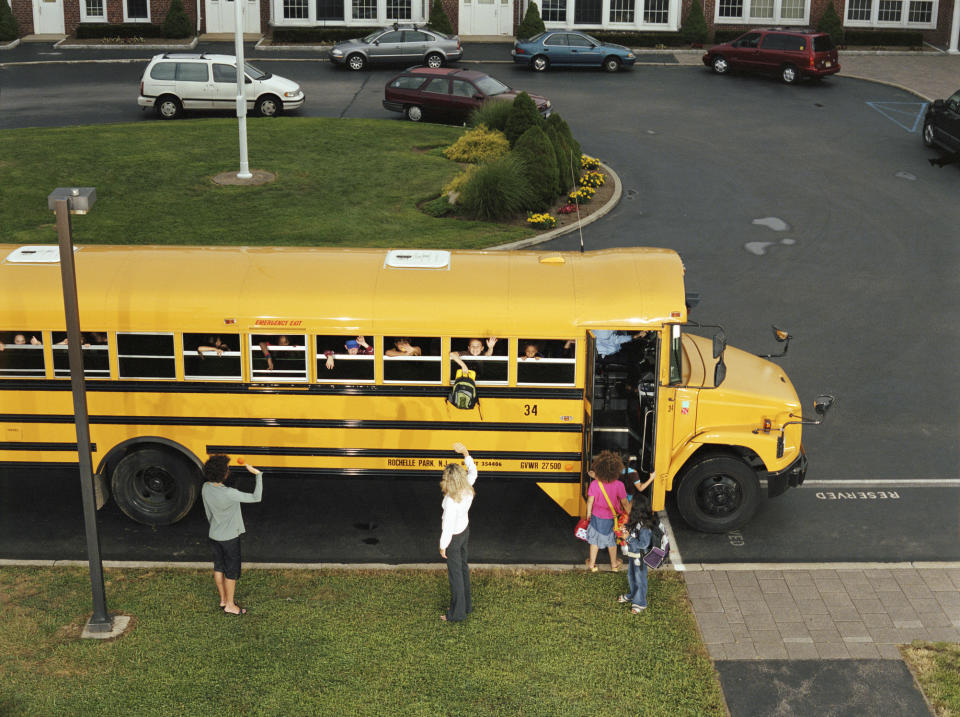 Schoolchildren waving from bus windows while adults greet them from the sidewalk