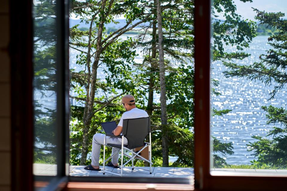 A man in a cap sits on a folding chair with a view of the lake and pine trees and works.