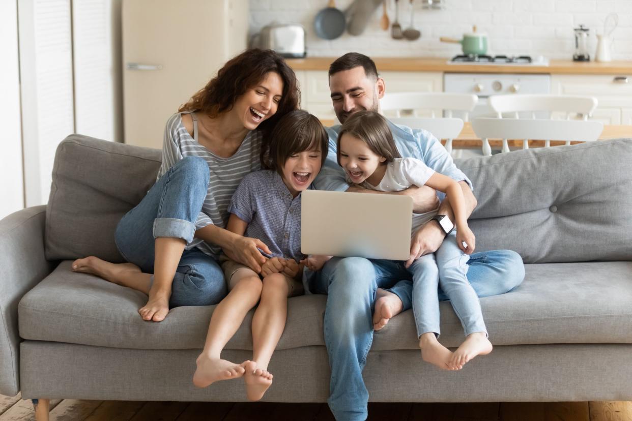 Overjoyed kids sitting on sofa with cheerful parents, watching funny video on computer. Happy married couple enjoying spending weekend time with small children, laughing looking at laptop screen.