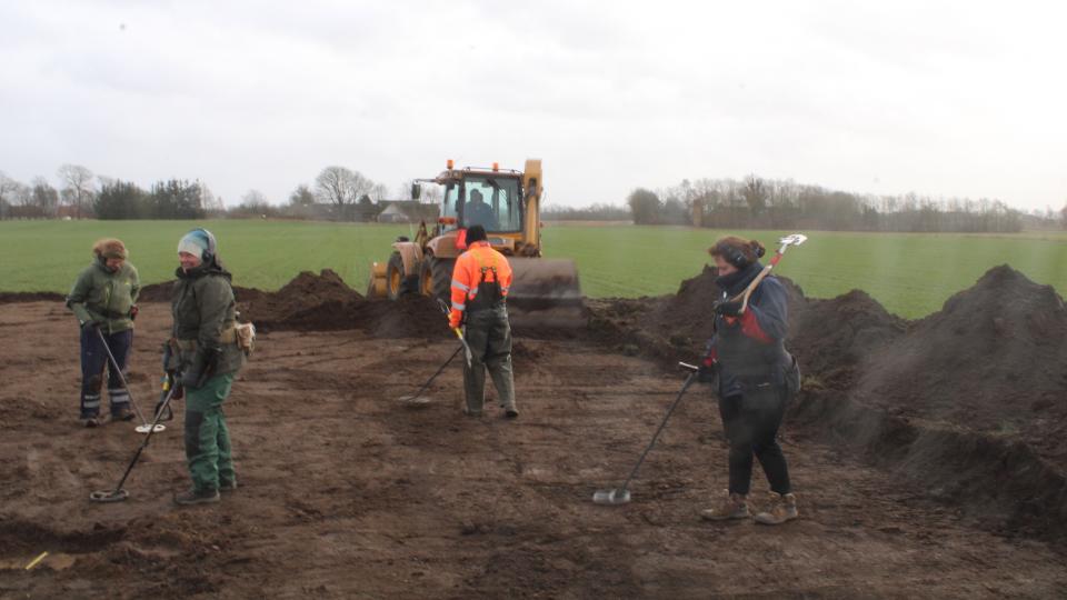 The location where the silver was found is very close to the site of Fyrkat, which was one of several distinctive ring forts built by Harald Bluetooth throughout Denmark. There are four people exploring a patch of upended dirt with metal detectors. There is a large yellow construction vehicle in the background.