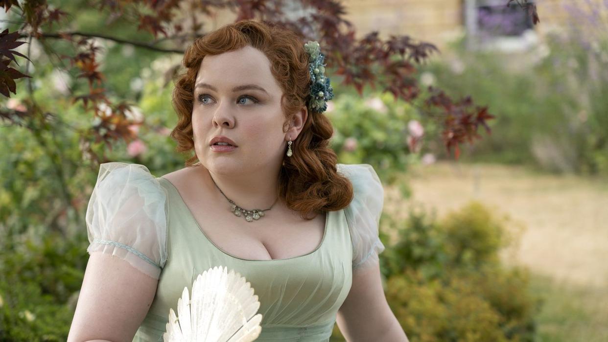 nicola coughlan as penelope featherington in bridgerton, she wears a green scoopneck dress and holds a fan in one hand while standing outside