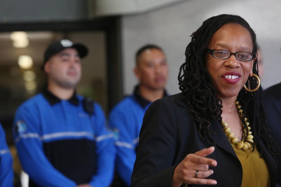 lateefah simon smiles and points with one hand, she wears a black suit jacket over an olive green top with a beaded necklace, hoop earrings, and glasses