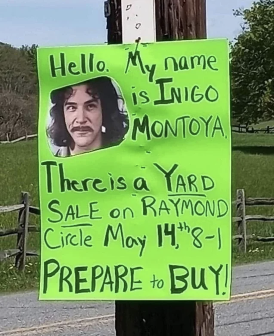 A sign on an outdoor pole — "Hello, my name is Inigo Montoya, there is a yard sale on Raymond Circle, May 14, 8–1, prepare to buy!" — with a photo of Mandy Patinkin as Inigo