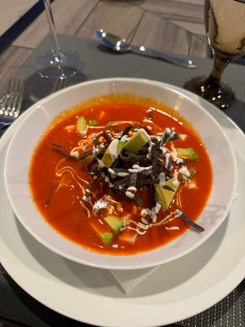 A bowl of vibrant soup garnished with avocado slices, tortilla strips, and drizzled with cream, placed on a neatly set table with cutlery and a wine glass