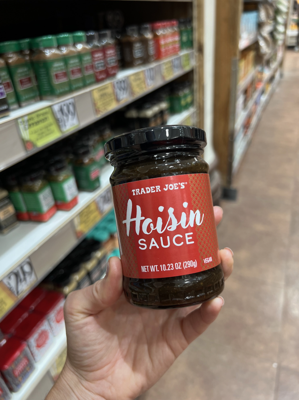 Hand holding a jar of Trader Joe's Hoisin Sauce in a grocery store aisle. Shelves stocked with various other sauces and spices are visible in the background