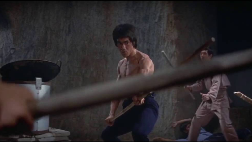 Bruce Lee stares down an enemy holding a staff.