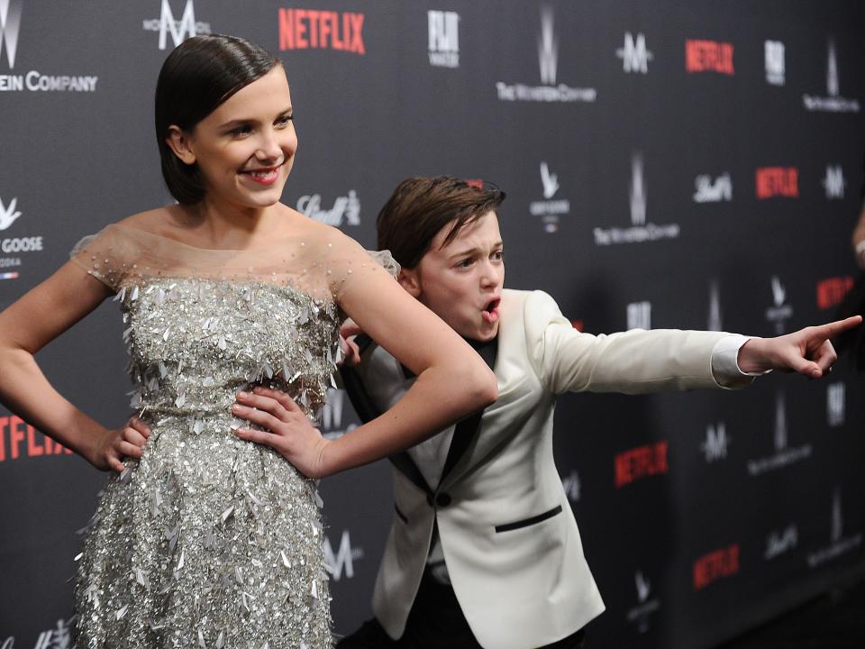 Millie Bobby brown and noah schnapp in 2017 at a golden globes afterparty red carpet. brown is in front, posing with her hands on her hips and wearing a sparkling silver dress. schnapp is behind her, posting with an excited expression on his face and pointing forward