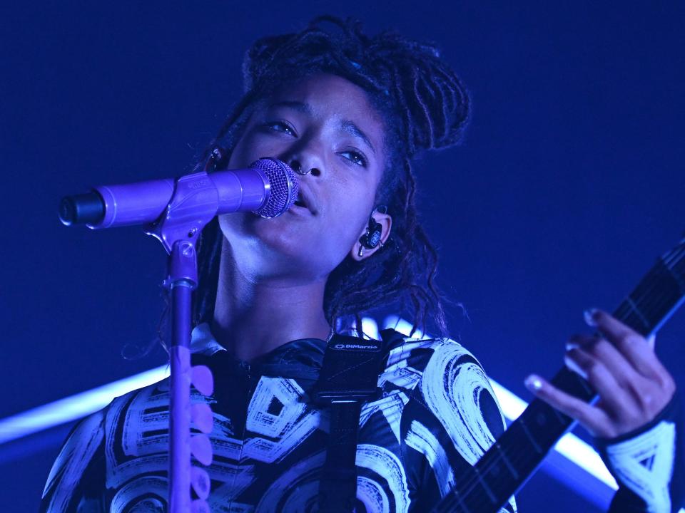 Willow Smith is performing on stage with guitar in hand. The stage lighting is blue and purple.