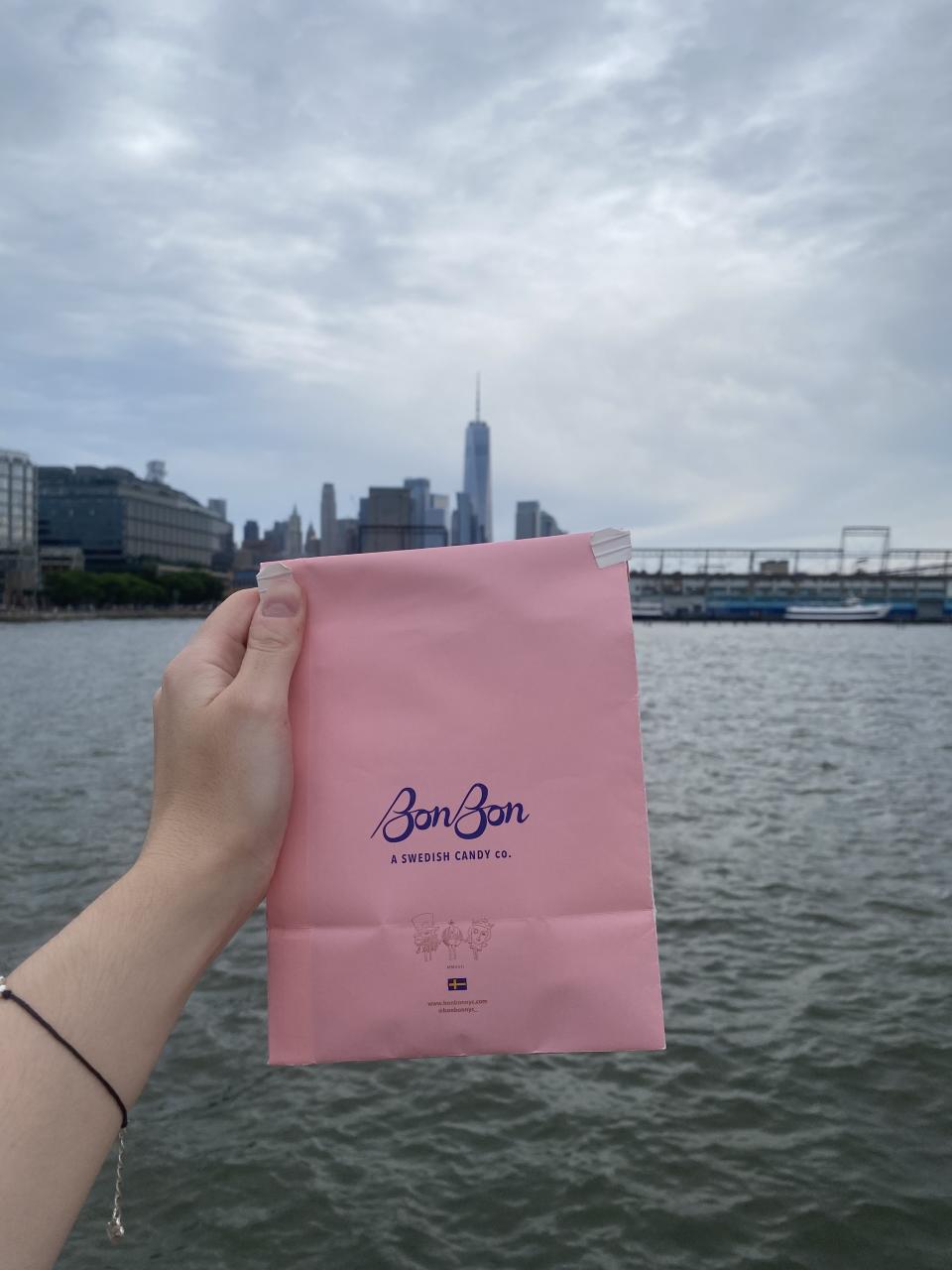 A hand holding a BonBon candy bag close to the water with buildings in the background. The bag says "BonBon, A Swedish Candy Co." and shows the brand's logo