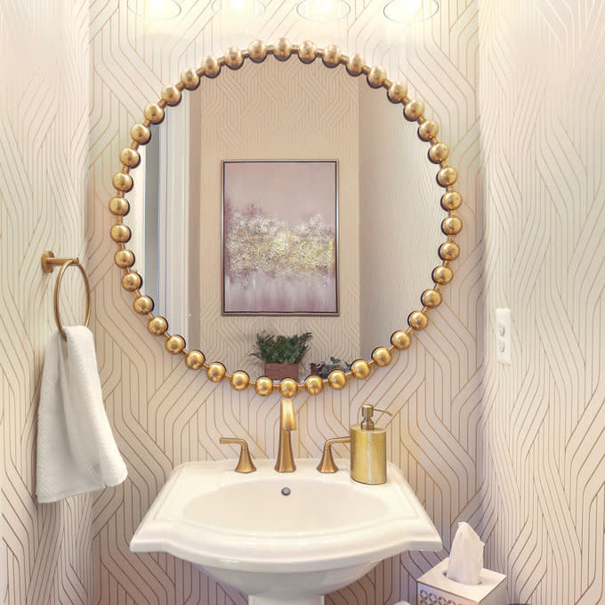 Bathroom scene with a round mirror framed by golden beads, a white pedestal sink, towel ring with white towel, soap dispenser, tissue box, and wall art