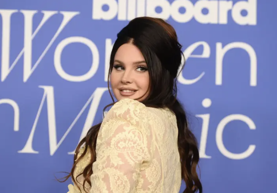 Lana del rey smiles in front of a lilac "women in music" sign at a press event