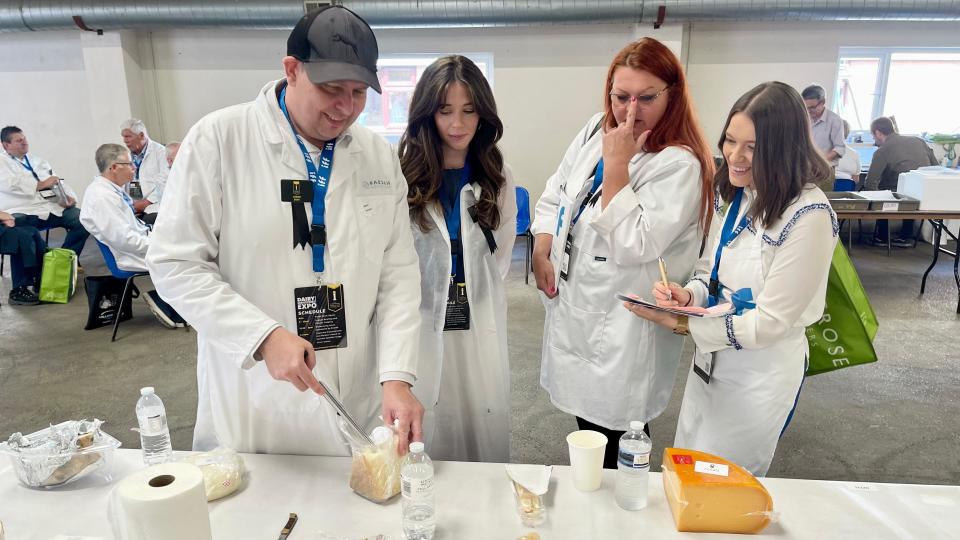 Four judges dressed in white coats inspect slabs of cheese. All are wearing white laboratory-style jackets with one man, on the left, wearing a cap and three women, all with long hair