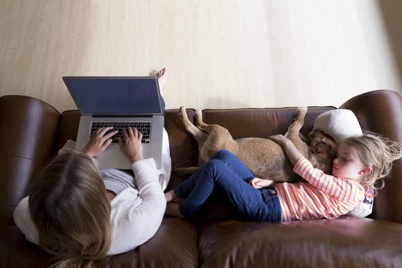 A woman types on a laptop next to a child cuddling a dog.