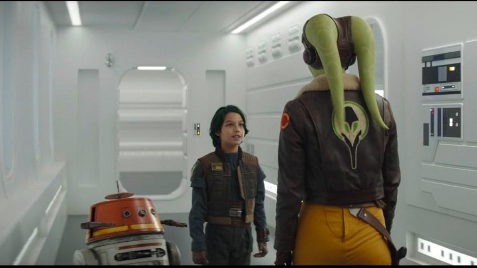 star wars ahsoka, jacen syndulla in a brown jacket with green hair speaks to green alien hera while standing next to chopper the droid