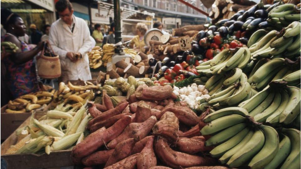 Customers weigh up the bananas, sweet potatoes or yams and peppers on sale on a fruit and vegetable market stall, Brixton, London, England circa 1969. (Photo by RDImages/Epics/Getty Images)