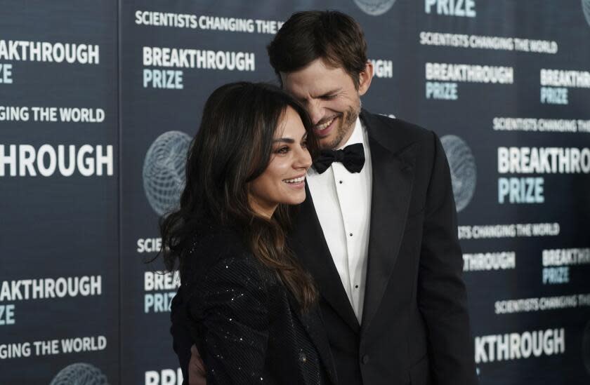 Ashton Kutcher smiling and laughing in a tuxedo while posing with a smiling Mila Kunis in a sparkly black dress.
